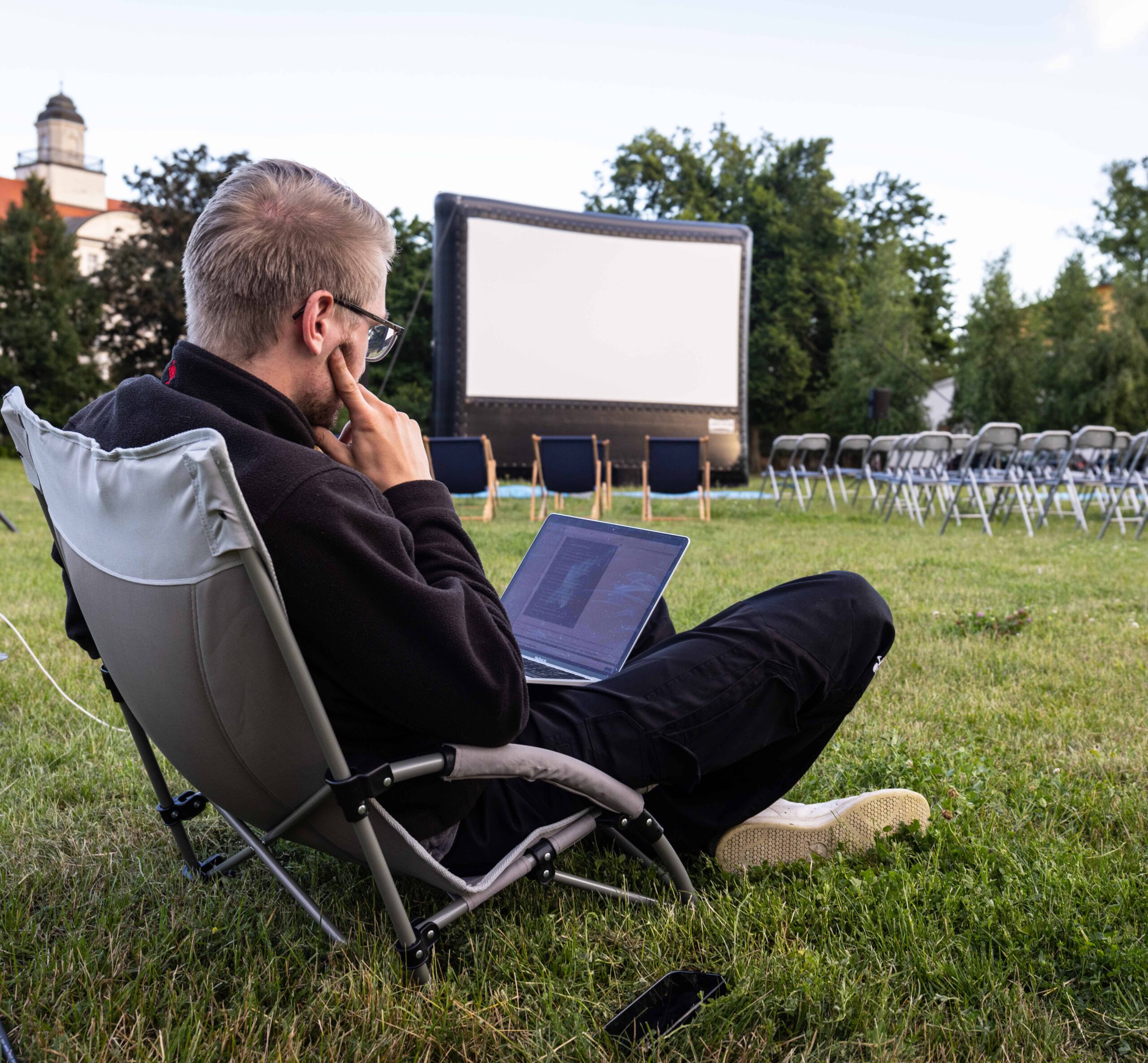 Ein Mitarbeiter sitzt auf einem Stuhl mit einem Laptop und macht die letzten technischen anpasungen, dahinter stehet die Open Air Kino Leinwand sowie die Stühle für den Besucher.