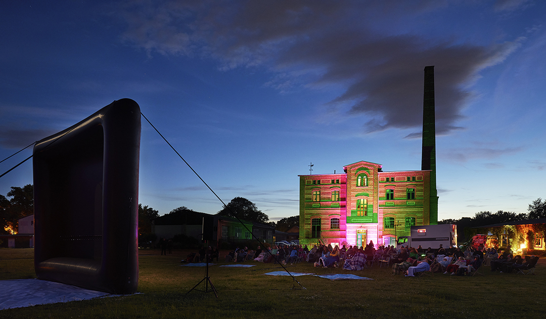 Freiluftkinoabend in Brandenburg. Das Projektionsfahrzeug ist geparkt und der Film läuft. Viele Besucher sitzen auf Stühlen und schauen gebannt zum Film.