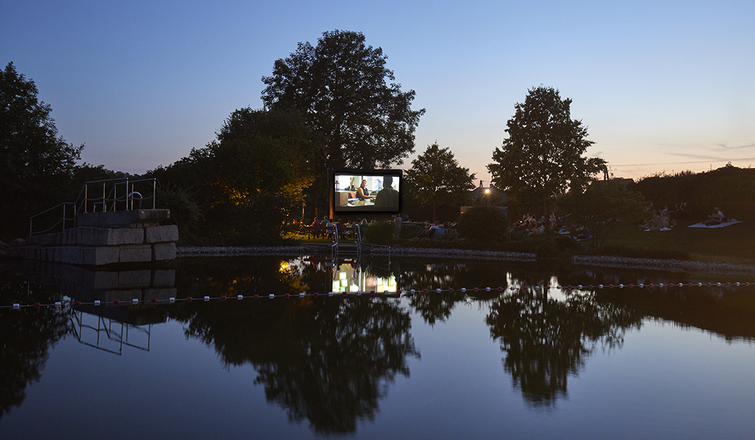 Freiluftkino im Freibad, das Bild auf der Leinwand spiegelt sich im Wasser. Es ist ein toller Sommerabend mit warmen Temperaturen